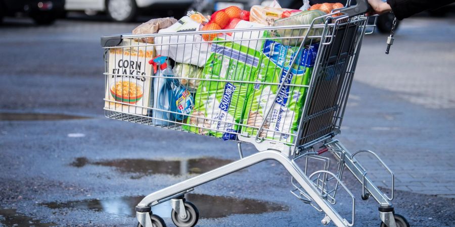 Ein Kunde schiebt einen bis zum Rand gefüllten Einkaufswagen über den Parkplatz eines Supermarktes.