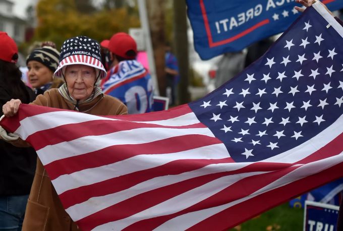 Eine Anhängerin vom US-Präsidenten Trump trägt einen «Keep-America-Great»-Hut und hält eine amerikanische, während sie am Rand einer Strasse, wartet. Wahlkampf in den USA