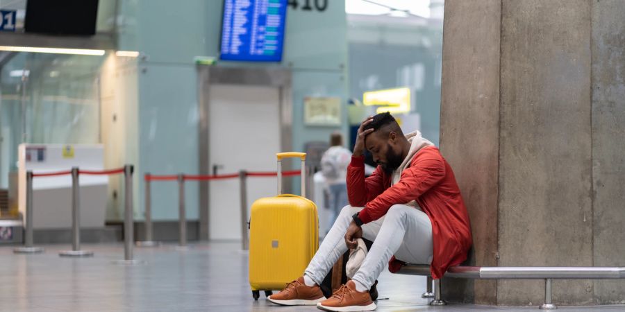 Schwarzer Mann wartet frustriert auf dem Boden am Flughafen mit einem gelben Koffer.