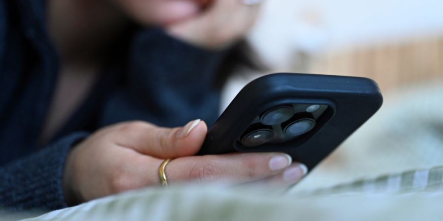 ILLUSTRATION - Eine junge Frau liegt mit dem Smartphone in der Hand auf dem Bett. Foto: Elisa Schu/dpa