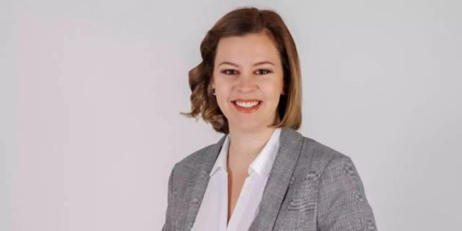 Frau mit schulterlangem braunen Haar, weisses Hemd und grauer Jacke