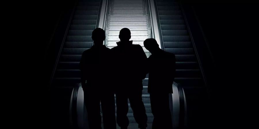 Drei Männer stehen im Dunkeln vor einer Treppe zwischen zwei Rolltreppen.