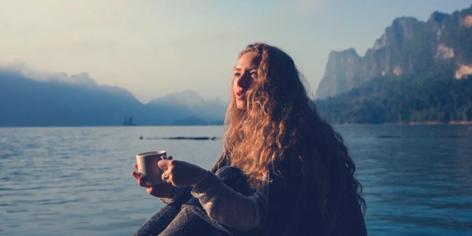 Alleine zu reisen ist eine gute Art, nach einer schmerzhaften Trennung wieder in seine Mitte zu kommen. Frau sitzt am See mit einer Tasse in der Hand.