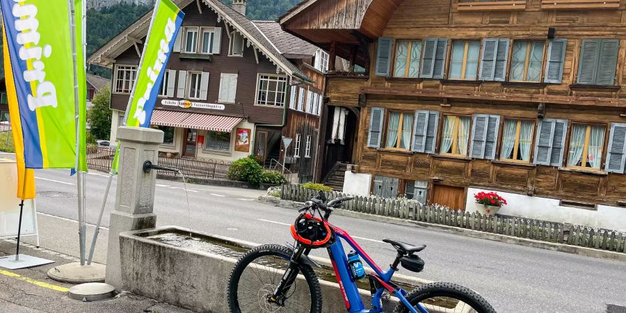 E-Mountainbike, Brunnen, Holzhäuser und die Fahnen von Post und Volg in Wimmis.