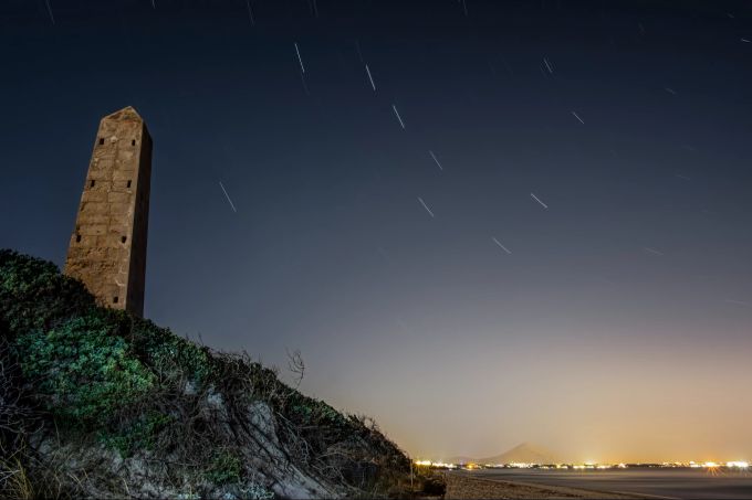 Nachtbild eines Strandes mit einem Steinmonolithen, Lichtern und Stadt im Hintergrund in Playa de Muro, Palma de Mallorca, Spanien Nachtbild Sterne Mallorca