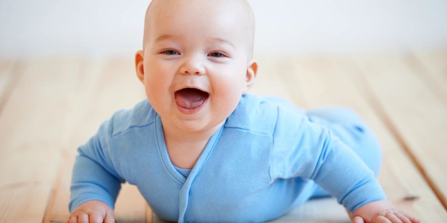 Baby lachend in blauem Onesie auf dem Boden liegend.