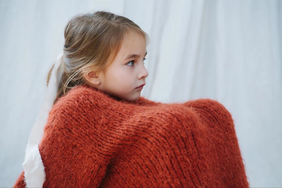 Kleines Mädchen mit Knien unter dem orangen Pullover wirkt traurig.