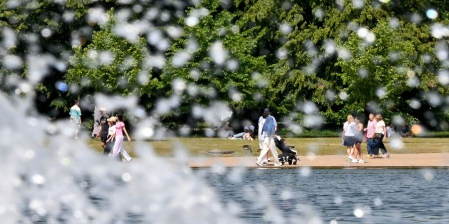 Menschen laufen bei sommerlichen Temperaturen durch die Kensington Gardens, während ein kleiner Springbrunnen Wasser versprüht. Foto: Anna Ross/dpa
