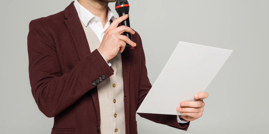 Mann mit Mikrofon in der Hand trägt bordeauxfarbenen Blazer zu weissem Hemd und Weste in Beige