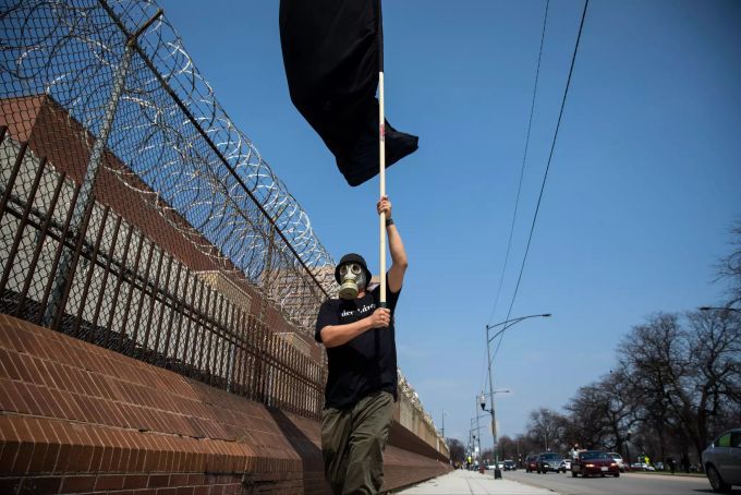 07.04.2020, USA, Chicago: Ein Mann mit Gasmaske schwenkt eine schwarze Fahne vor dem Cook County Gefängnis. Inmitten der Coronavirus-Pandemie fahren Demonstranten um das Gefängnis und das Leighton-Strafgericht und fordern mit Hupen und Sprechchören die «Massenfreilassung» der Gefangenen. Foto: Ashlee Rezin Garcia/Chicago Sun-Times/dpa +++ dpa-Bildfunk +++ Coronavirus - USA