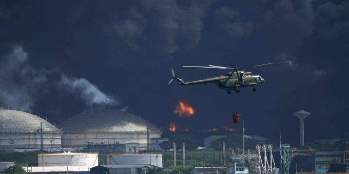 Ein mit Wasser beladener Hubschrauber fliegt über die Supertanker-Basis Matanzas, von der dunkler Rauch und Flammen aufsteigen. Ein mit Wasser beladener Hubschrauber fliegt über die Supertanker-Basis Matanzas, von der dunkler Rauch und Flammen aufsteigen.