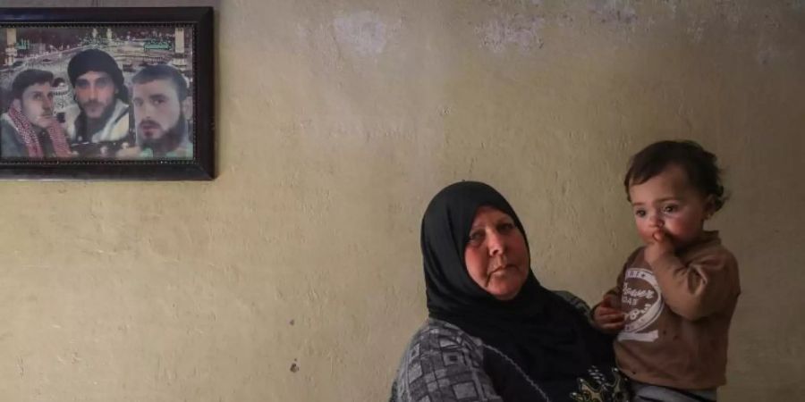 Die 63-jährige Syrerin Samiha Seri hält in Idlib eines ihrer 16 verwaisten Enkelkinder auf dem Arm. Hinter ihr an der Wand ein Bild mit Fotos von dreien ihrer Söhne. Foto: Anas Alkharboutli/dpa