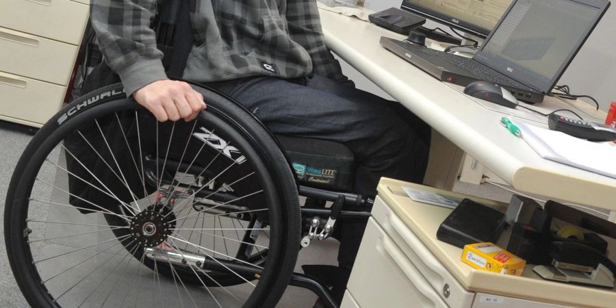 Ein Mann sitzt in einem Rollstuhl an seinem Arbeitsplatz am Schreibtisch.