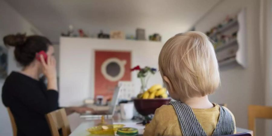 Eine Frau sitzt im Homeoffice an ihrem Laptop und telefoniert, während ihr Kind neben ihr in einem Kinderstuhl am Tisch sitzt. Foto: Christian Beutler/KEYSTONE/dpa