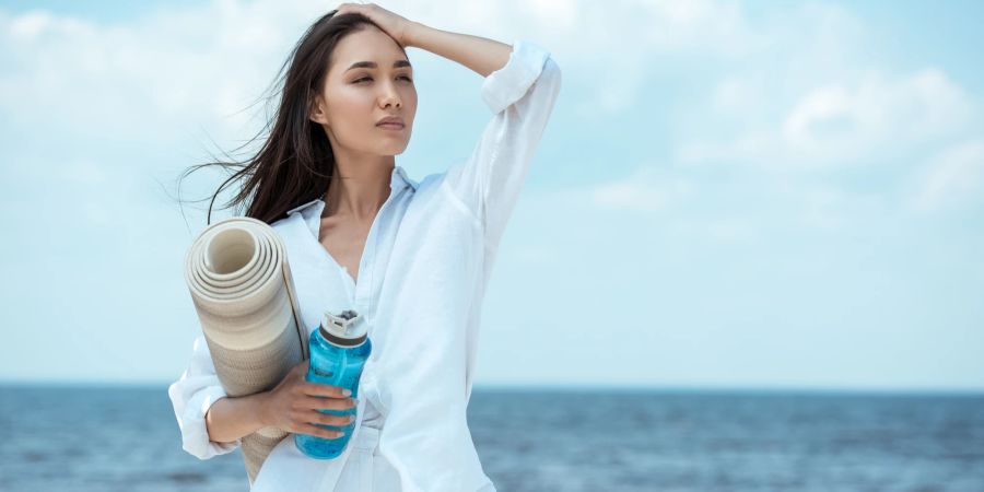 Asiatische Frau am Meer mit Wasserflasche und Yogamatte.