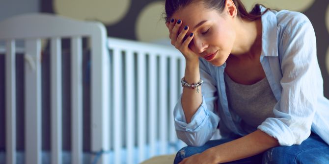 Überforderte, nachdenkliche Frau sitzt vor einem Kinderbett