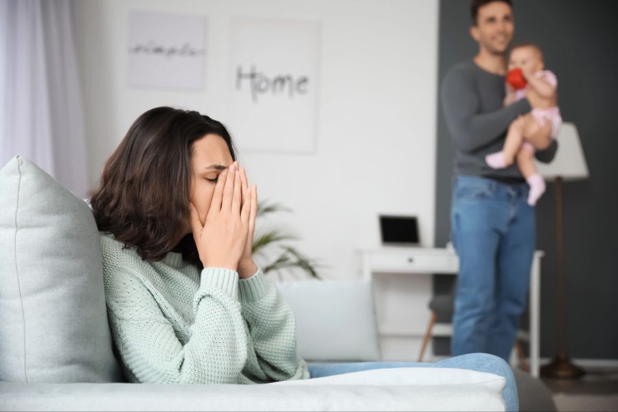 Erschöpfte Mutter auf einem Sofa, Mann mit Kind im Hintergrund