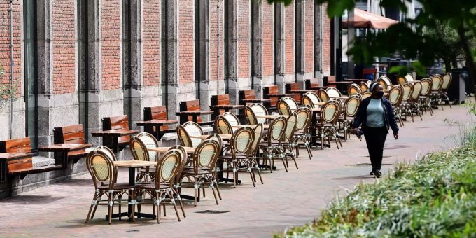 Eine Frau mit schwarzem Mundschutz geht bei sonnigem Wetter an den leeren Tischen und Stühlen eines Restaurants vorbei. Gastronomie