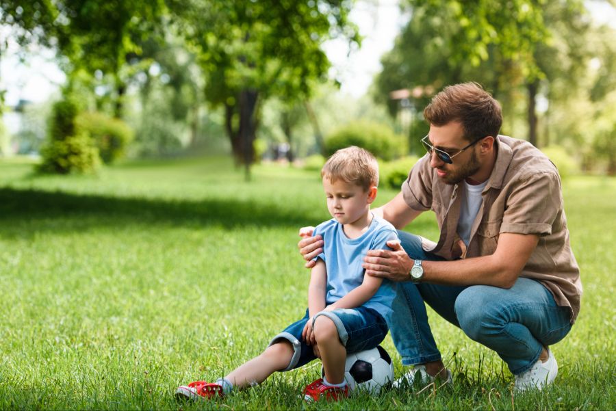 Vater tröstet kleinen Jungen, der auf einem Fussball sitzt