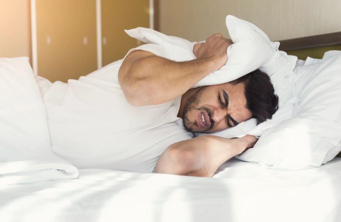 Laute Zimmernachbarn? Dann hilft nur: Ohren zuhalten oder gleich nach dem Upgrade fragen. Bärtiger Mann liegt im Bett und hält sich mit Kissen die Ohren zu.