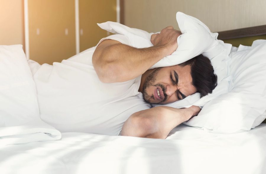 Bärtiger Mann liegt im Bett und hält sich mit Kissen die Ohren zu.