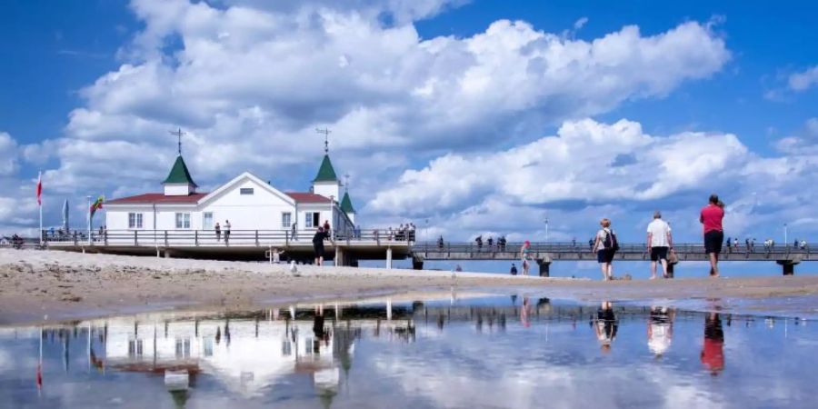 Urlauber spiegeln sich bei einem Strandspaziergang vor der Seebrücke in einer Wasserfläche. Foto: Jens Büttner/dpa