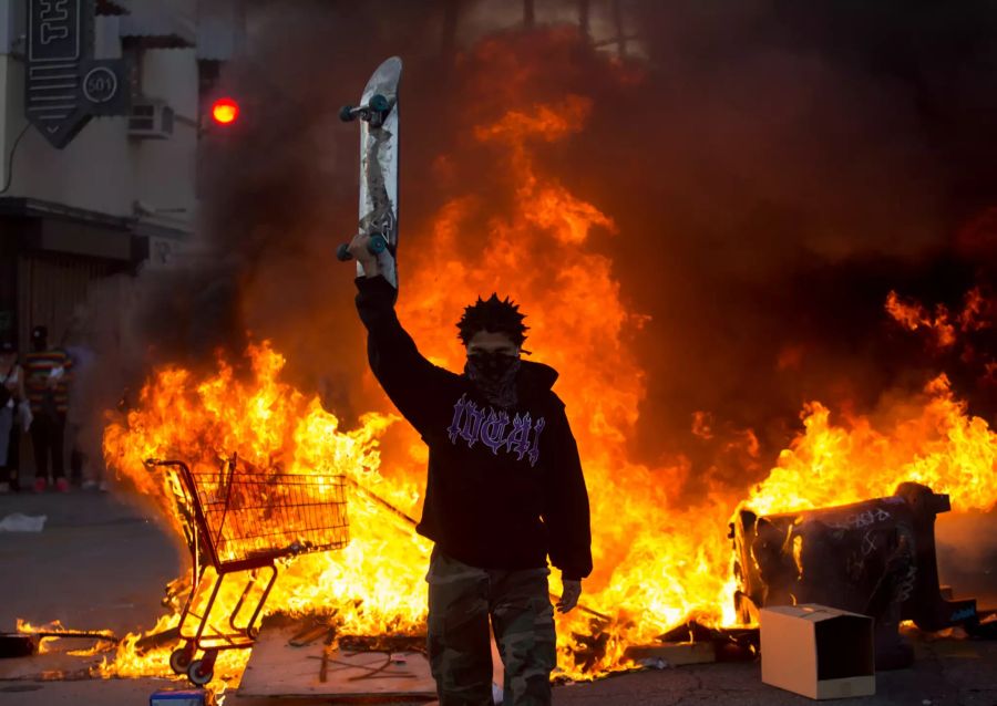 Ein Demonstrant hält ein Skateboard in die Höhe, während hinter ihm ein Einkaufswagen auf der Strasse brennt.