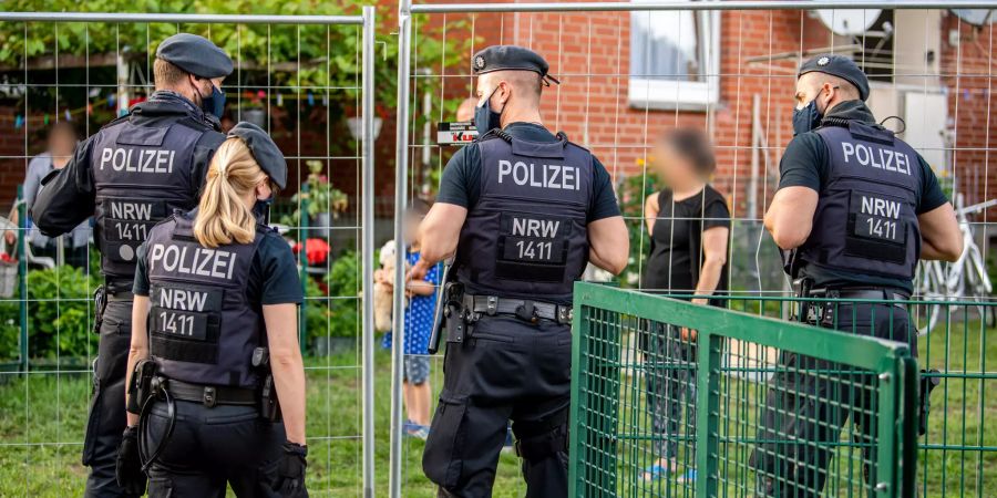 Polizisten sprechen mit Bewohnern, die hinter einem Bauzaun unter Quarantäne stehen. Die Stadt Verl hat nach positiven Corona-Tests bei zahlreichen Tönnies-Mitarbeitern im Stadtteil Sürenheide eine Quarantäne eingerichtet.