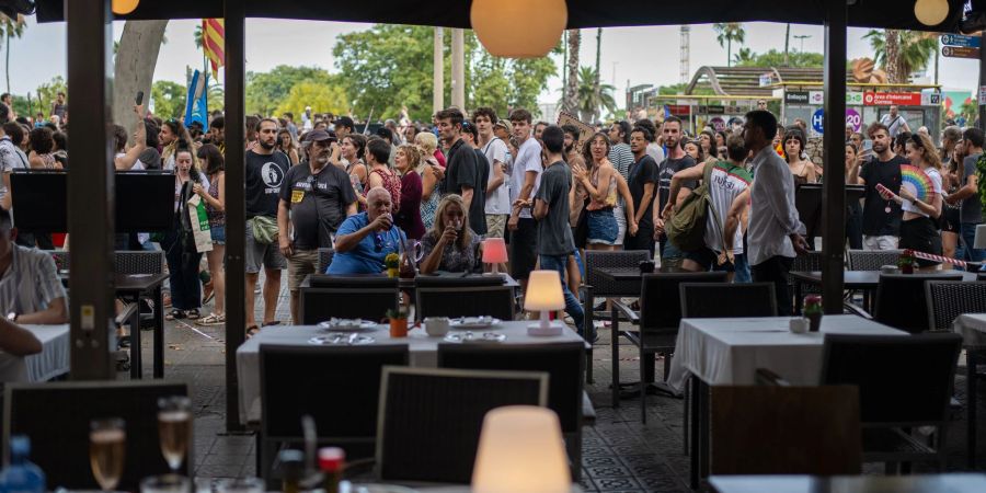 Ein Mann und eine Frau auf der Terrasse eines Restaurants in Barcelona lassen sich von vorbeiziehenden Demonstranten gegen den Massentourismus offenbar nicht aus der Ruhe bringen.