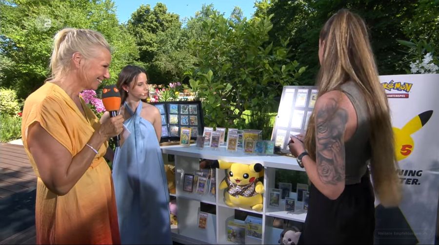 Andrea Kiewel schaut sich mit zwei Pokémon-Sammlerinnen ein mehrsprachiges Pikachu-Kartenset an.