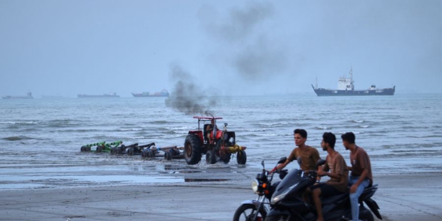 ARCHIV - Junge Männer fahren mit Motorrädern am Ufer entlang, während ein Traktor Geräte aus dem Wasser zieht und Handelsschiffe in der Straße von Hormuz vor Anker liegen. Foto: Ra...