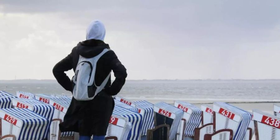 Grau und windig auf Norderney: Eine Frau steht warm angezogen auf der Strandpromenade und schaut über die verwaisten Strandkörbe hinweg auf die Nordsee. Foto: Volker Bartels