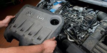 Ein KFZ-Servicetechniker in einer Autowerkstatt hält die Abdeckung vor einem 2.0l TDI Dieselmotor vom Typ EA189 .
