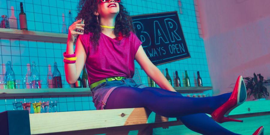 Frau in blauen Strumpfhosen, pinken Pumps und roter Sonnenbrille sitzt Shot-trinkend auf einer Bar.