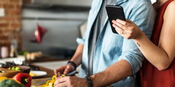 Mann und Frau bei der Essenszubereitung schauen auf Smartphone