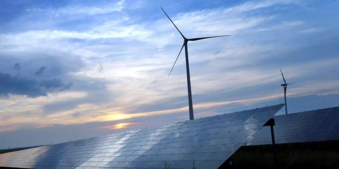 Der mit Wolken überzogene Abendhimmel spiegelt sich auf den Solarzellen einer vor Windrädern stehenden Solarkraftanlage. (Symbolbild) Anleihe