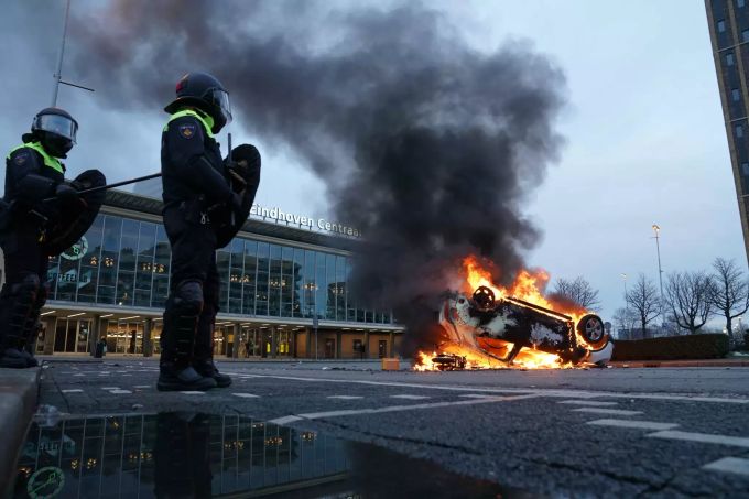Flammen schlagen aus einem Auto, das vor dem Bahnhof Eindhoven auf dem Kopf liegt. Mehrere hundert Menschen haben vor dem Bahnhof gegen die aktuelle Corona-Politik demonstriert. Coronavirus
