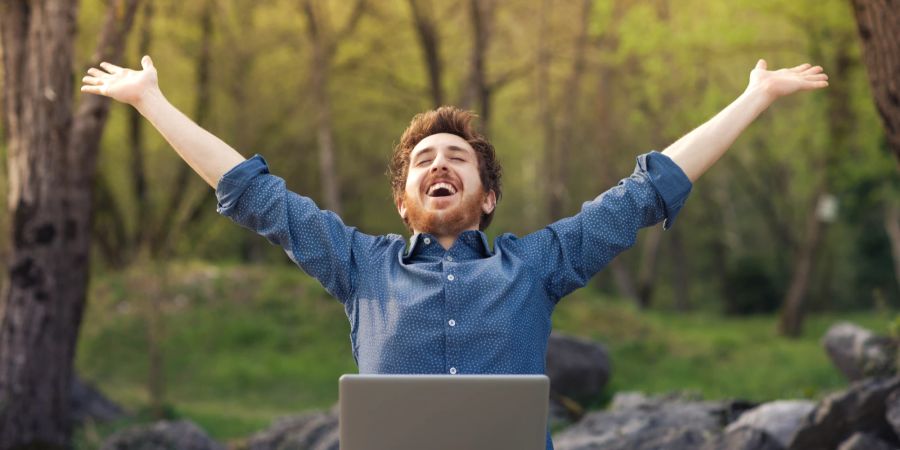 Mann im Freien am Laptop wirft die Arme freudig in den Himmel.