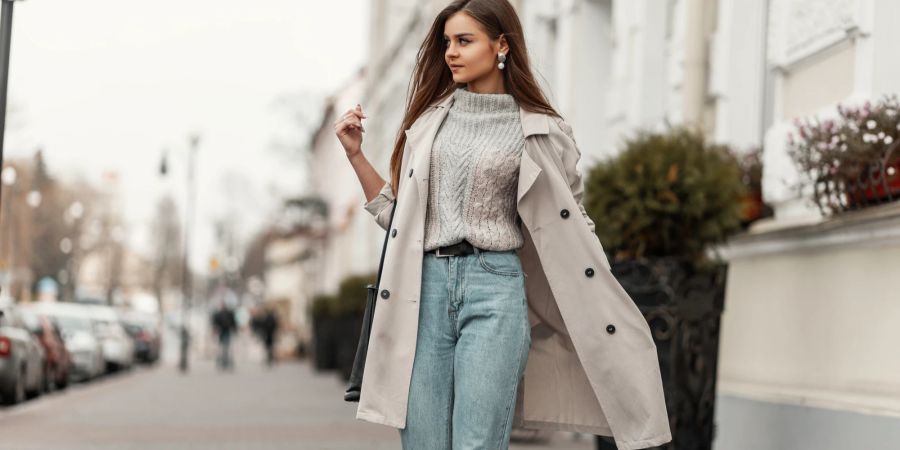 Frau im modischen Trenchcoat in einem stylischen Strickpullover mit schwarzer Ledertasche spaziert durch die Stadt.