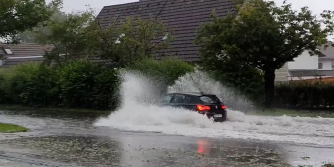 Ein Auto fährt durch das stehende Wasser auf einer Strasse im Ort und schiebt eine Bugwelle vor sich her. In Teilen Bayerns gab es am Sonntag heftige Unwetter. Unwetter in Teilen Bayerns