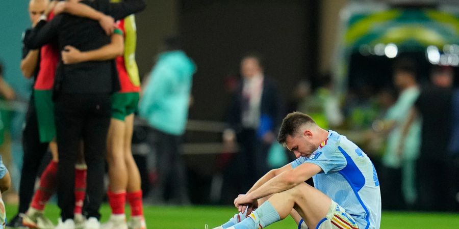 Spaniens Aymeric Laporte sitzt nach der Niederlage auf dem Spielfeld, während Marokkos Spieler im Hintergrund über ihren Sieg im Elfmeterschiessen jubeln.