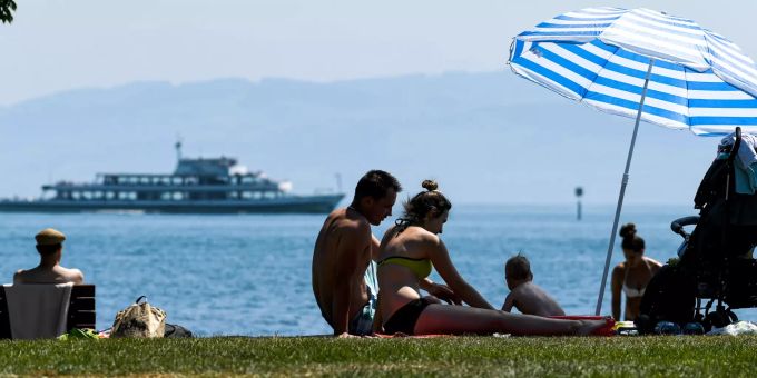 Ein Paar sitzt mit seinem kleinen Sohn am Ufer des Bodensees in der Sonne, während im Hintergrund ein Passagierschiff vorbeifährt. Ferienpark am Bodensee