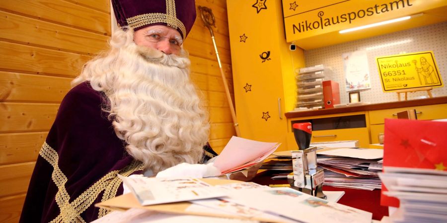 Ein als Nikolaus verkleideter Mann sitzt im Nikolauspostamt in St. Nikolaus vor vielen Briefen.