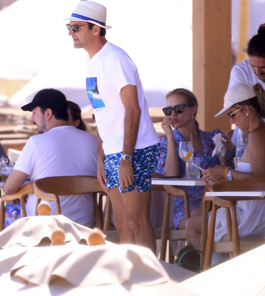 Roger Federer im Sommer-Look: Der Maestro trägt bunte Badehose, weisses Shirt und Panamahut. Gattin Mirka Federer (rechts mit Mütze) tippt auf ihrem Handy.