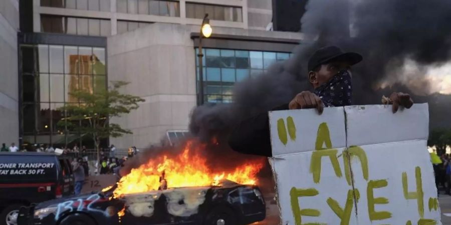 Ein Demonstrant hält bei einer Protestaktion in der Nähe eines brennenden Polizeiautos ein Schild mit dem Anfang eines Zitats aus der Bibel «Auge um Auge». Foto: Ben Gray/Atlanta Journal-Constitution/AP/dpa