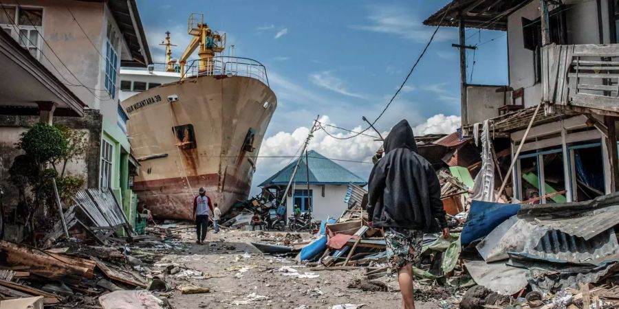 Menschen gehen eine Strasse entlang in der Nähe des Ortes Palu in Indonesien, wo ein Schiff während des Tsunamis an Land gespült wurde.