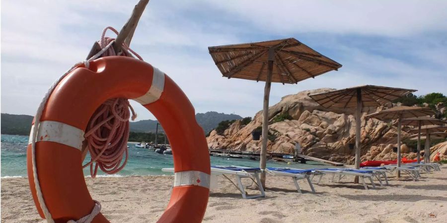 Ein Rettungsring steht neben Sonnenliegen und Schirmen am Sandstrand von Capriccioli (ITA).