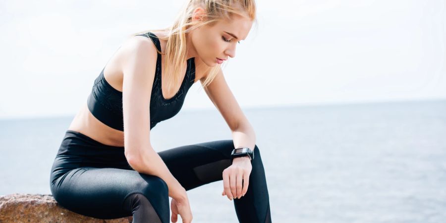 Eine Frau sitzt in Sportkleidung auf einem Stein und schaut auf ihre Smart Watch.