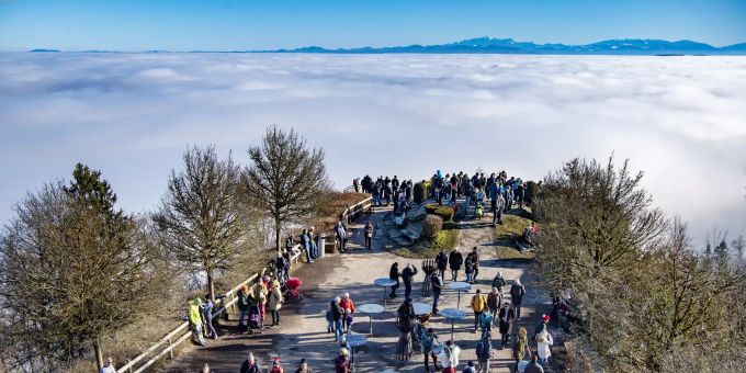 Menschen stehen auf einer Aussichtsplattform auf dem Uetliberg, während sich im Hintergrund eine dichte Nebeldecke erstreckt. Nebel über dem Züricher Uetliberg