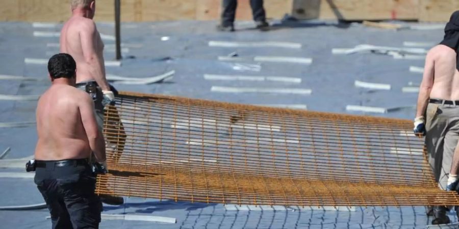 Arbeiter mit freiem Oberkörper tragen auf einer Baustelle ein Stahlgitter. Foto: Ole Spata